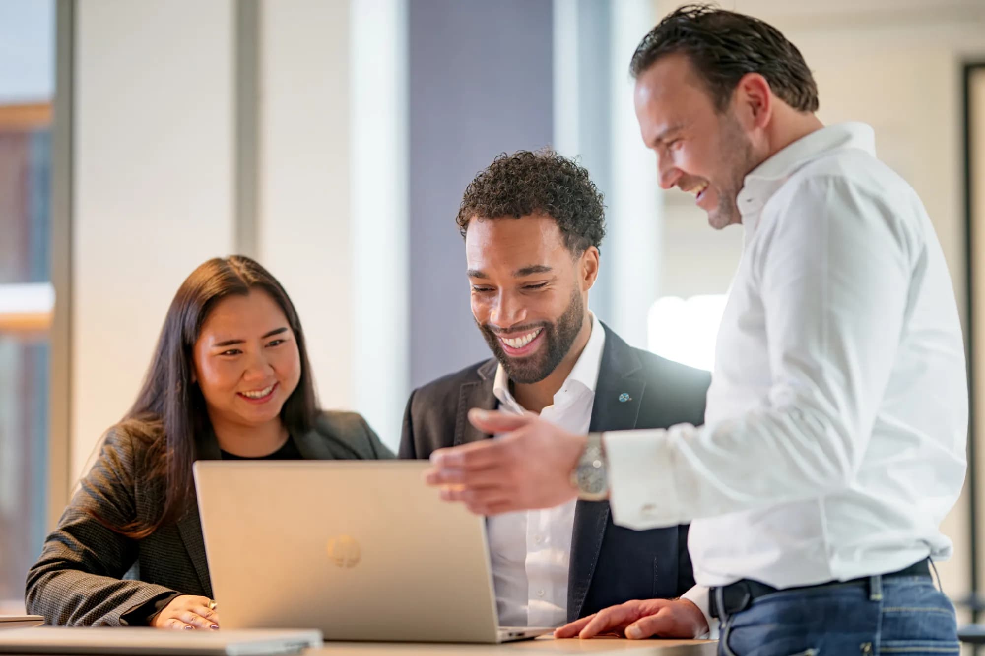 HR-tech oplossingen: de drijvende kracht achter toekomstbestendig talentmanagement. Drie collega's kijken lachend naar een laptop tijdens een meeting op kantoor.